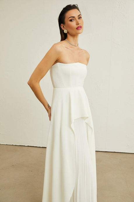 A woman wearing a white offshoulder maxi dress with pleated details standing against a plain background