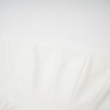 A woman wearing a white offshoulder maxi dress with pleated details standing against a plain background