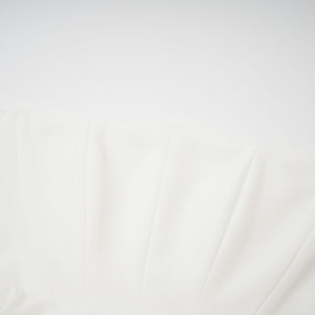 A woman wearing a white offshoulder maxi dress with pleated details standing against a plain background