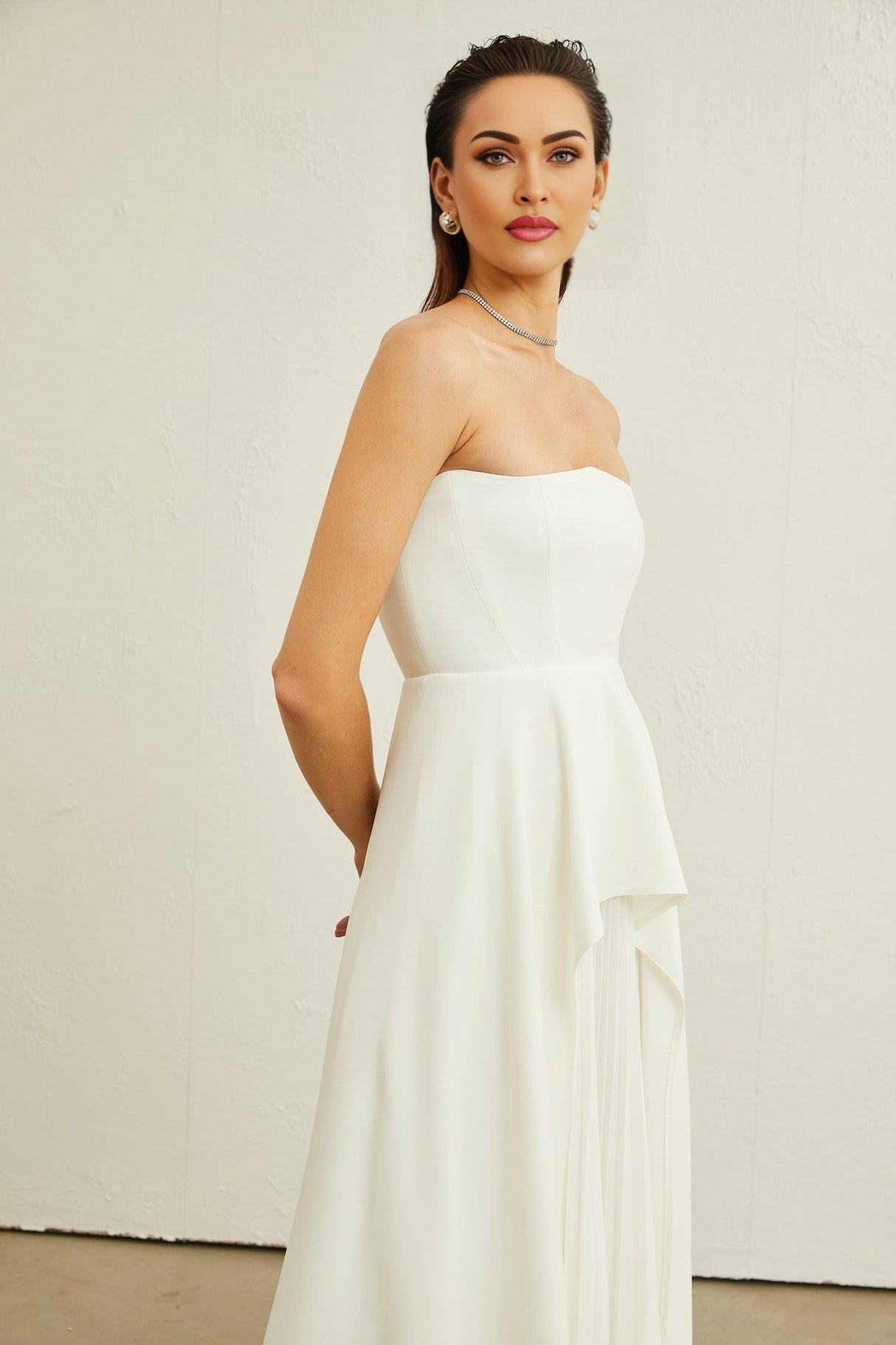 A woman wearing a white offshoulder maxi dress with pleated details standing against a plain background