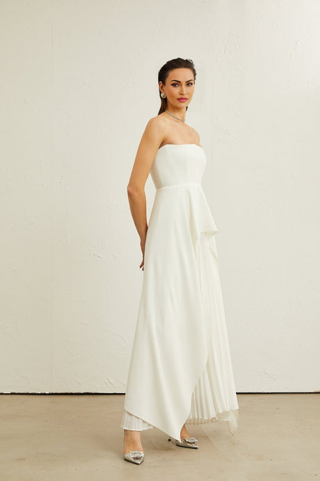 A woman wearing a white pleated offshoulder maxi dress with a flowing skirt standing against a plain background