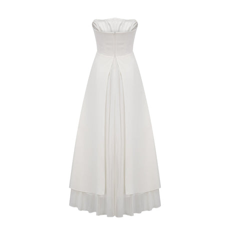 A woman wearing a white pleated offshoulder maxi dress with a flowing skirt standing against a plain background