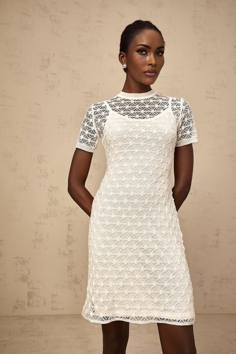A woman wearing a white semisheer knit mini dress with a closefitting silhouette standing against a plain background