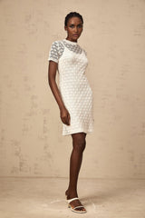 A woman wearing a white semisheer knit mini dress with short sleeves standing against a plain background
