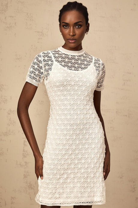 A woman wearing a white semisheer knit mini dress with short sleeves standing against a plain background
