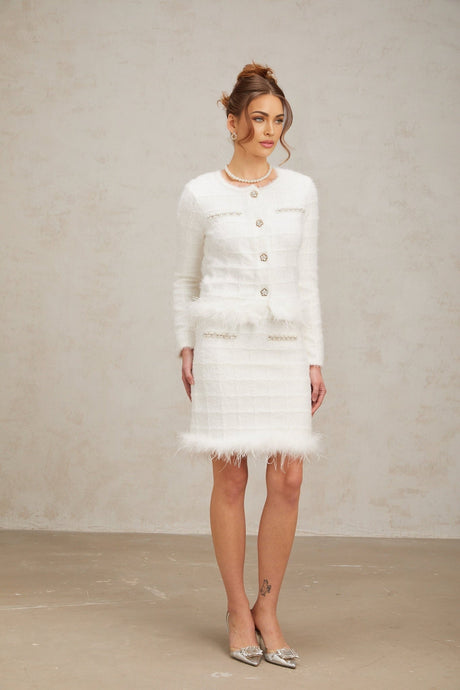A woman wearing a white waffle knit cardigan and matching skirt set with feather details standing against a plain background