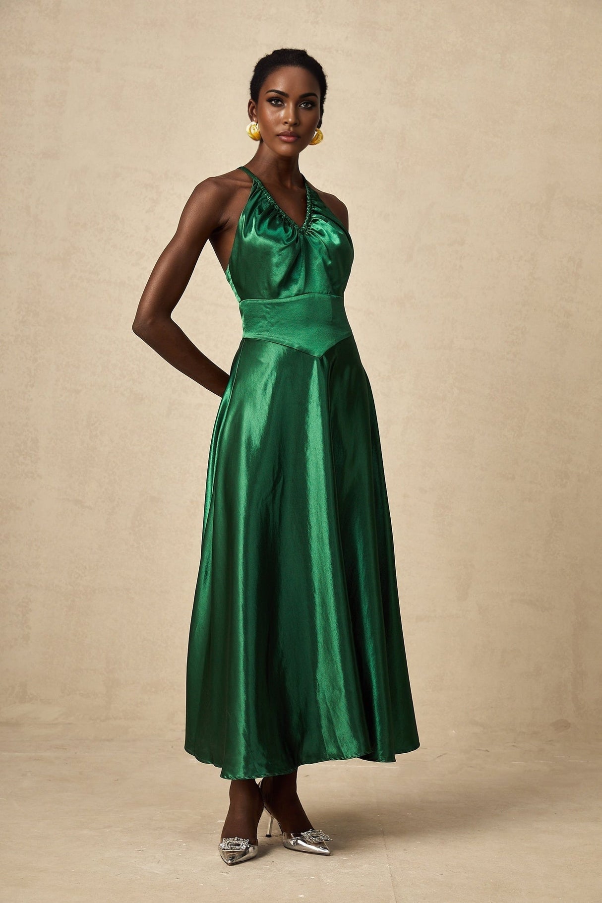A woman wearing an emerald green satin midi dress with an open back design standing against a plain background