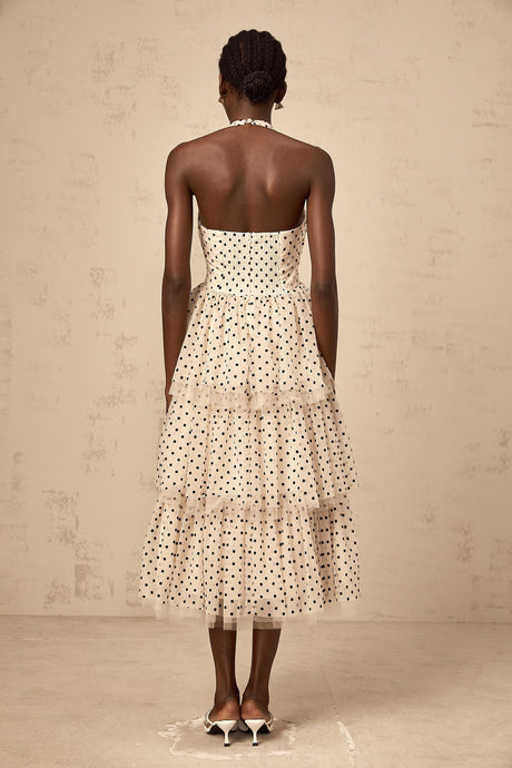 A woman wearing an ivory colored midi dress with polka dots and tiered layers standing in a neutral background