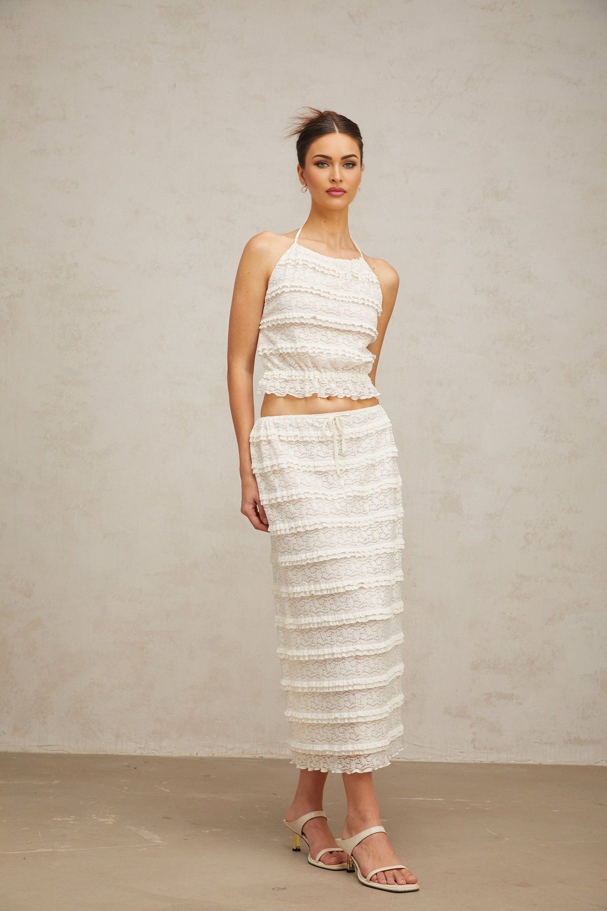 A woman wearing an off white floral lace matching set consisting of a ruffled top and skirt standing against a plain background