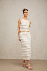 A woman wearing an off white floral lace matching set consisting of a ruffled top and skirt standing against a plain background