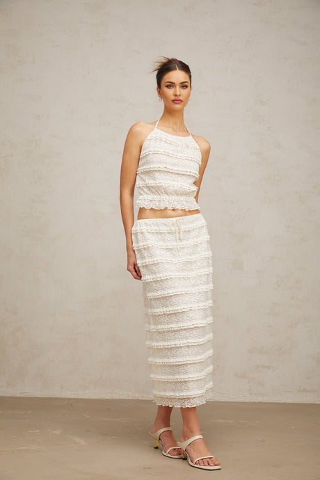 A woman wearing an off white floral lace matching set consisting of a ruffled top and skirt standing against a plain background