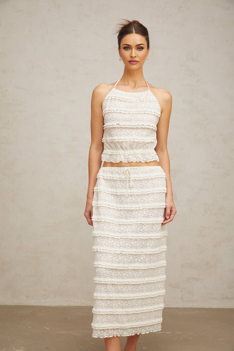 A woman wearing an off white floral lace matching set consisting of a ruffled top and skirt standing against a plain background
