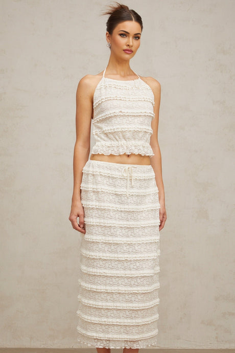 A woman wearing an off white floral lace matching set consisting of a ruffled top and skirt The outfit has delicate lace details and a feminine ruffled design