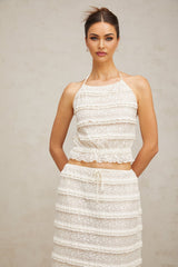 A woman wearing an off white floral lace matching set consisting of a ruffled top and skirt The set features delicate lace patterns and ruffled details creating a romantic and feminine look