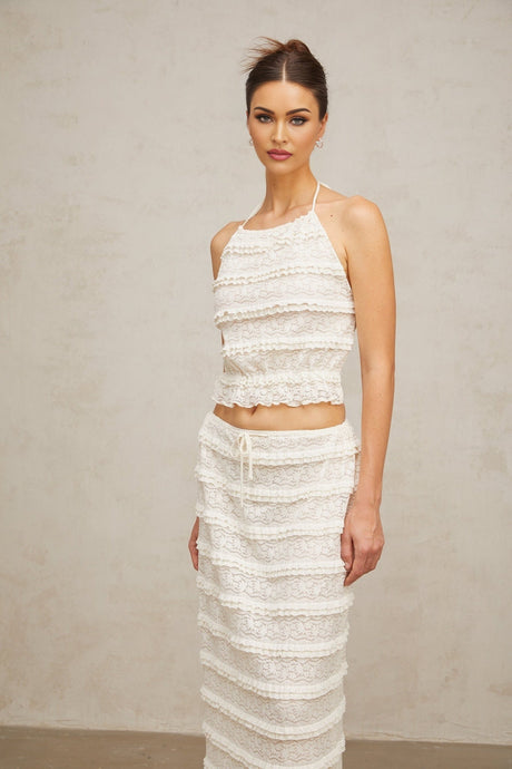 A woman wearing an off white matching set consisting of a floral lace top with ruffles and a coordinating skirt The ensemble has a romantic and delicate appearance