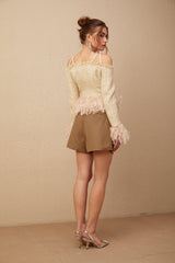 A woman wearing an offshoulder tweed top with feather trim around the neckline The top is light colored and has a textured appearance The model has her arms raised showing the offshoulder design