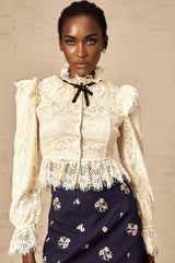 A woman wearing an offwhite blouse with floral lace detailing and a bowknot accent at the neckline