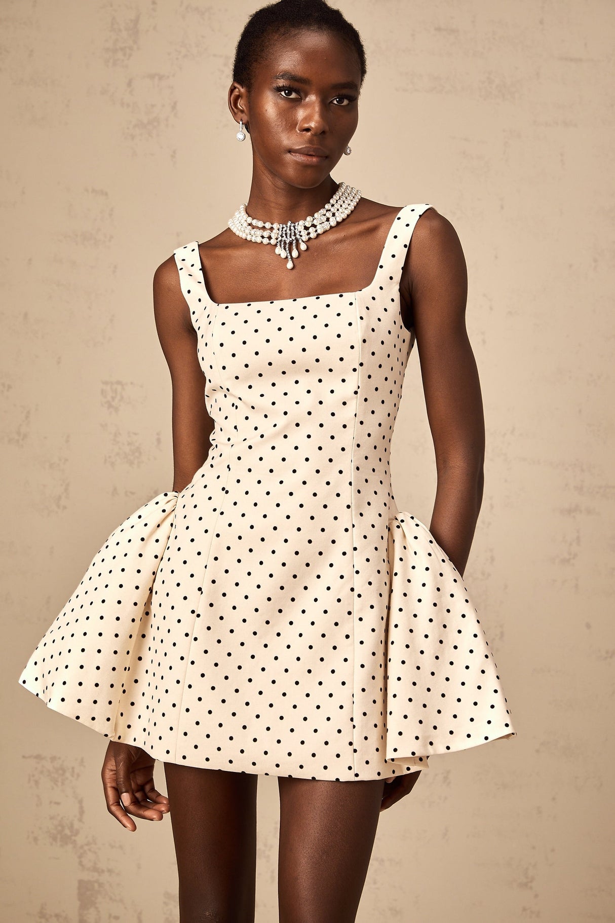 A young woman wearing a beige mini dress with white polka dots and puff sleeves standing against a neutral background