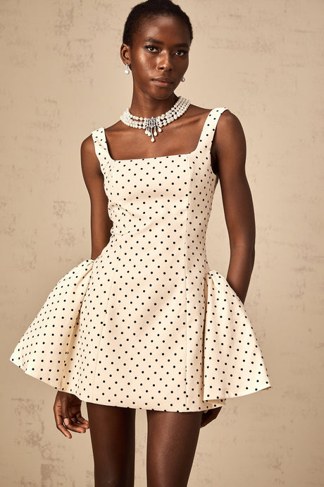 A young woman wearing a beige mini dress with white polka dots and puff sleeves standing against a neutral background