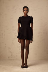 A young woman wearing a black mini dress with an asymmetric neckline and ruffle details posing against a plain background