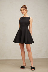 A young woman wearing a black sleeveless mini dress with a flared skirt standing against a plain background