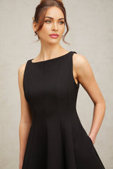 A young woman wearing a black sleeveless mini dress with a flared skirt standing in a neutral background