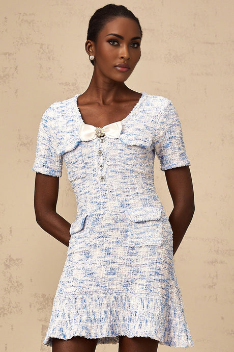 A young woman wearing a blue and white checked knit mini dress with short sleeves and a fitted silhouette standing against a plain background
