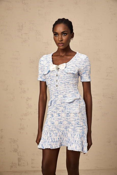 A young woman wearing a blue and white checked knit mini dress with short sleeves standing against a plain background