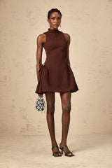 A young woman wearing a brown mini dress with an asymmetric neckline and puffed sleeves standing against a plain background