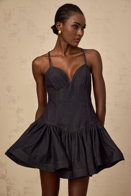 A young woman wearing a dark blue denim mini dress with a flounce detail standing against a plain background