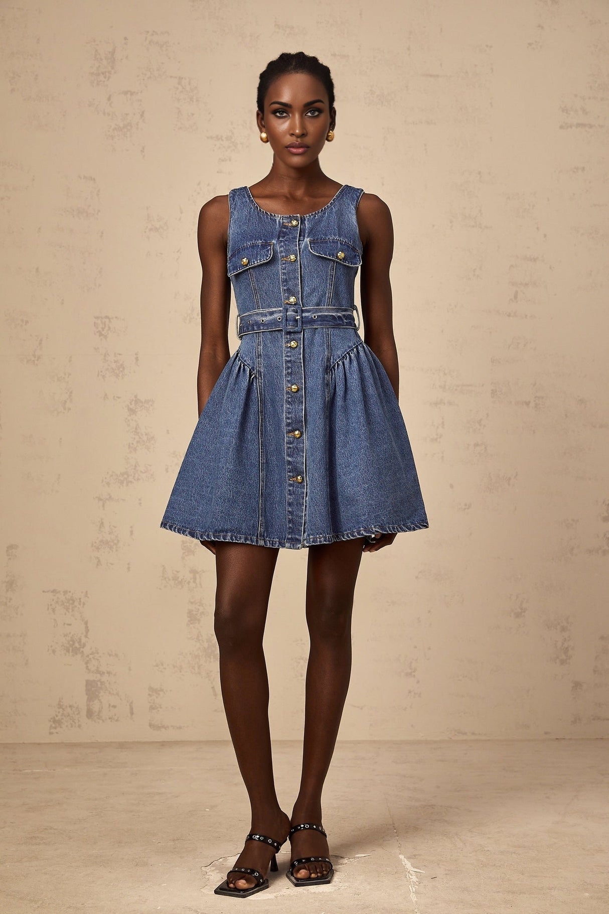 A young woman wearing a light blue denim mini dress with short sleeves and a belted waist standing against a plain background