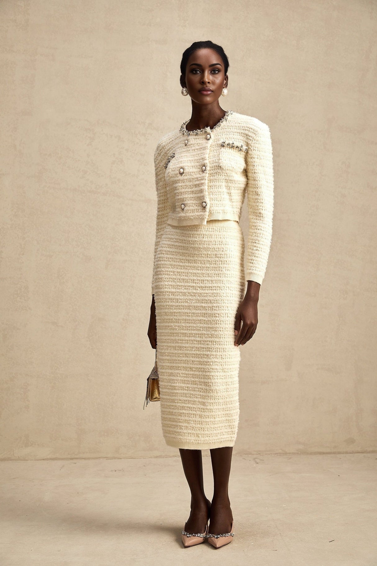 A young woman wearing a matching knit cardigan and skirt set with embellishments The outfit is light colored and features intricate detailing on both pieces