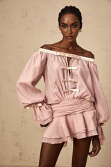 A young woman wearing a pink offshoulder mini dress with a bow detail and ruffled hem standing against a plain background