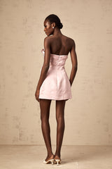 A young woman wearing a short pink Aline dress with a large bowknot detail at the waist posing against a plain background