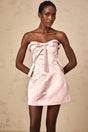 A young woman wearing a short pink dress with a bow at the waist and a flared skirt standing against a plain background