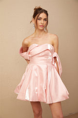 A young woman wearing a short pink offshoulder dress with puffed sleeves made of satin fabric standing against a plain background
