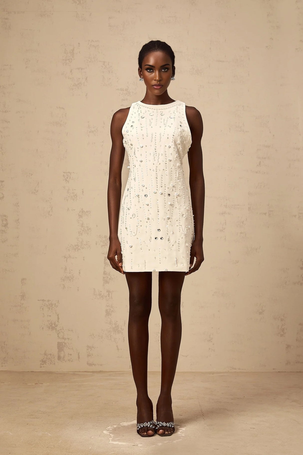 A young woman wearing a short white Aline dress with embellishments standing against a plain background