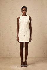 A young woman wearing a short white Aline dress with embellishments standing against a plain background