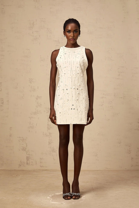 A young woman wearing a short white Aline dress with embellishments standing against a plain background