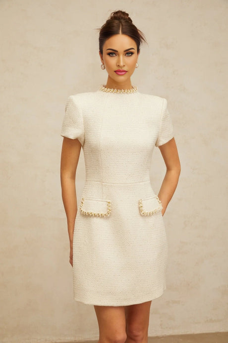 A young woman wearing a short white tweed dress with decorative embellishments standing against a plain background