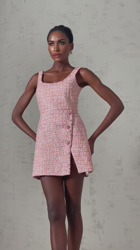 A young woman wearing a sleeveless pink tweed mini dress with embellishments standing against a plain background