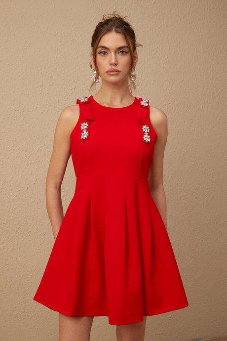 A young woman wearing a sleeveless red mini dress with a pleated skirt and a large bow detail at the neckline