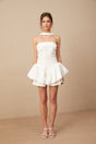 A young woman wearing a white halterneck mini dress with ruffled details standing against a plain background