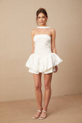 A young woman wearing a white halterneck mini dress with ruffled details standing against a plain background