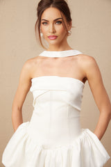 A young woman wearing a white halterneck mini dress with ruffled details standing against a plain background