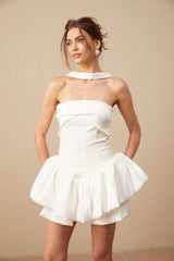 A young woman wearing a white halterneck mini dress with ruffled details standing against a plain background