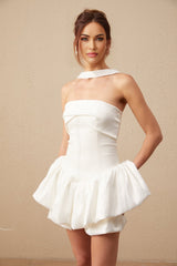 A young woman wearing a white halterneck mini dress with ruffled details standing against a plain background
