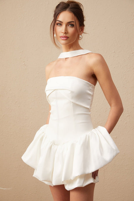 A young woman wearing a white halterneck mini dress with ruffled details standing against a plain background
