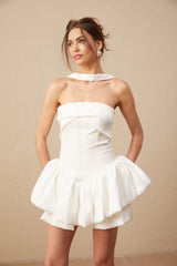 A young woman wearing a white halterneck mini dress with ruffled details standing against a plain background