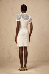 A young woman wearing a white semisheer knit mini dress standing in a relaxed pose against a plain background